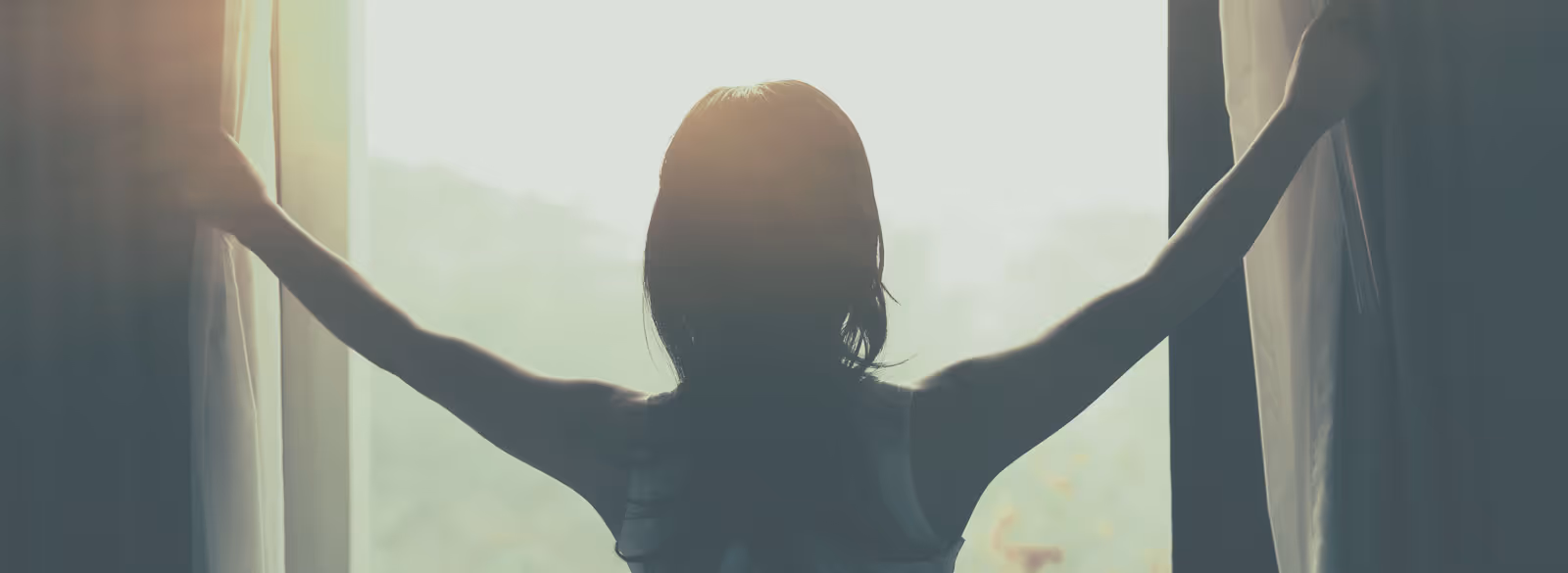 Silhouette of a person opening curtains with bright sunlight streaming in through the window.