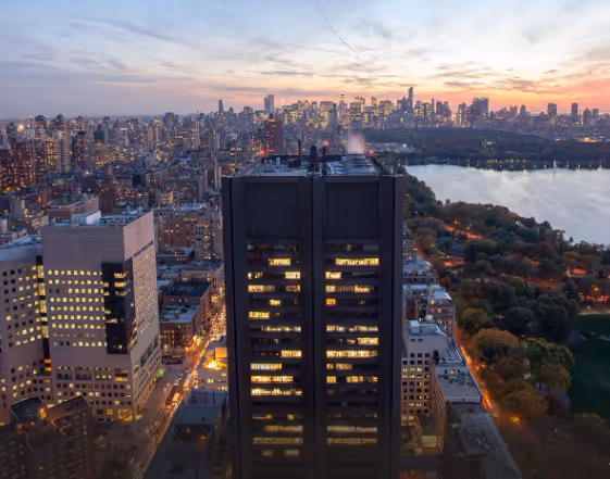 Aerial view of a city skyline at dusk with illuminated tall buildings, a large park, and a winding river.