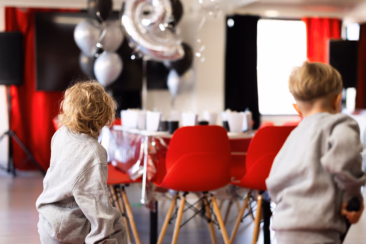 Kaksi lasta katsoo koristeltua juhlapöytää, jossa on punaisia tuoleja ja hopeisia ilmapalloja numerolla 3.