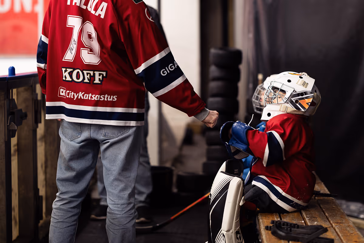 Lapsi jääkiekkomaalivahti istuu penkillä haarniskassa ja tekee nyrkkitervehdyksen aikuisen kanssa, joka seisoo vieressä punaisessa pelipaidassa.