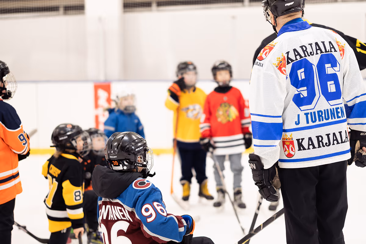 Jääkiekkovalmentaja ja joukko lapsia jäähallissa, lapset pukeutuneina eri joukkueiden peliasuihin ja kypäröihin.