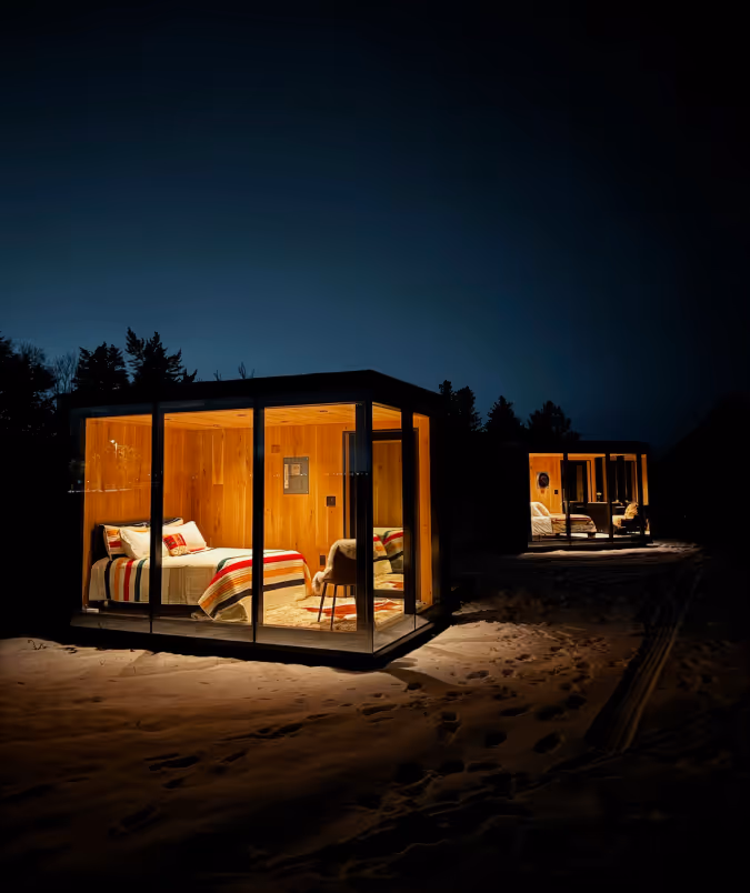 Two illuminated glass cabins with wooden interiors in a snowy landscape at night.