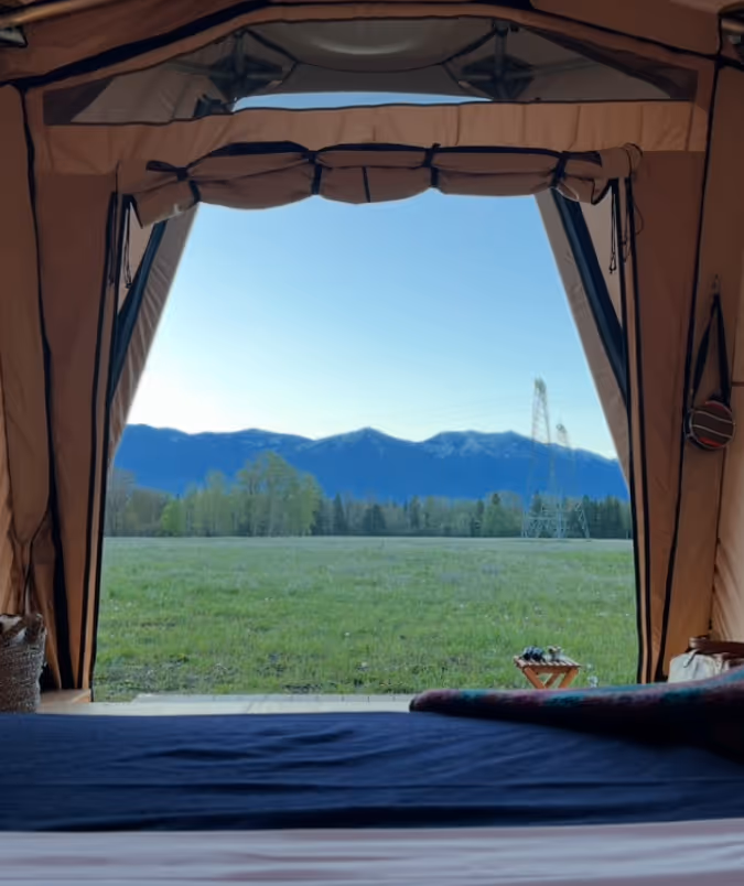 View of green field and distant mountains seen through the open entrance of a tent with a bed inside.