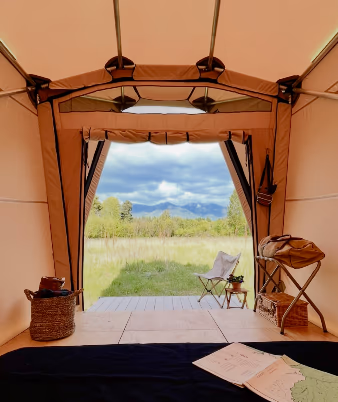 View from inside a spacious tent showing grassy field, trees, and mountains under a cloudy sky.