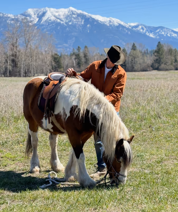Person in a cowboy hat and brown jacket adjusting the saddle of a brown and white horse grazing in a grassy field with snowy mountains in the background.