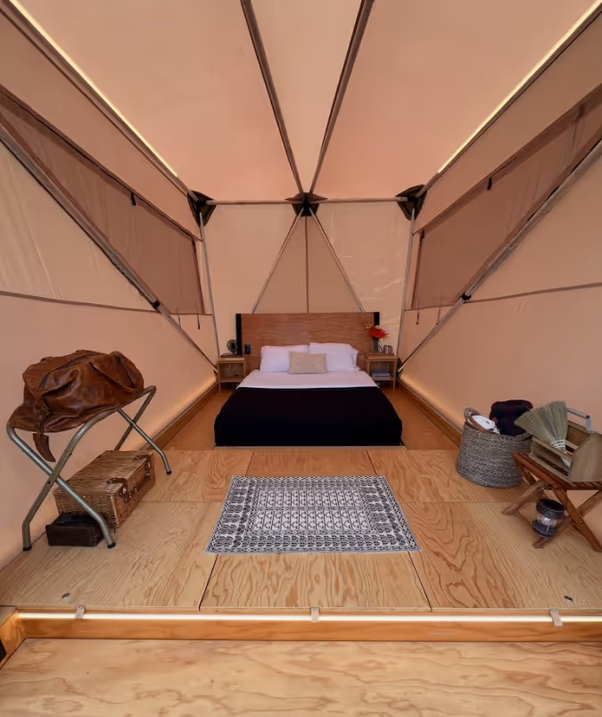 Interior of a glamping tent with a bed, wooden floor, small rug, luggage rack with a brown leather bag, and woven baskets.