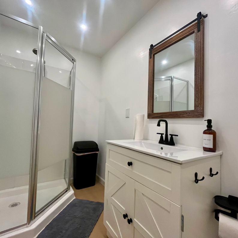 Modern bathroom with a white vanity, black faucet, wooden framed mirror, glass shower, and a black trash can.