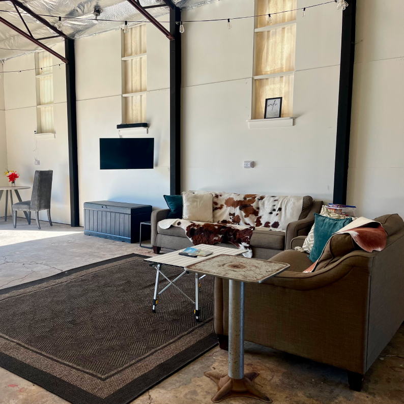 Industrial-style living area with two brown sofas, cowhide throws, a folding table, a dark rug, a wall-mounted TV, and a small table with a chair and flowers.