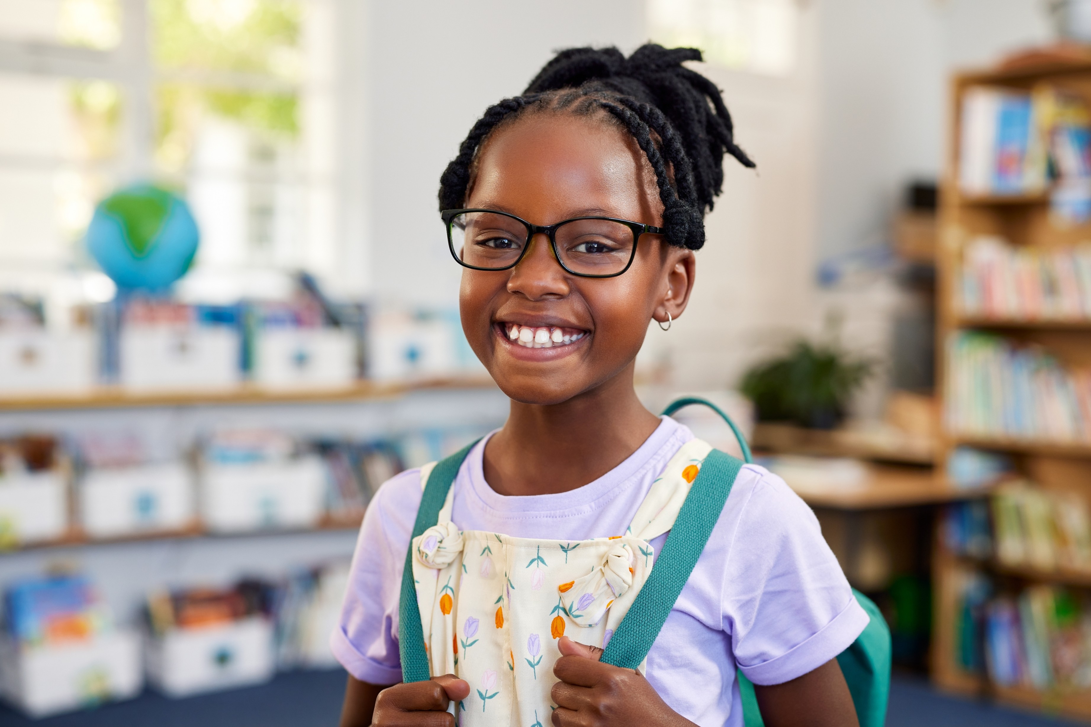 A smiling schoolgirl wearing glasses and a backpack in a classroom.