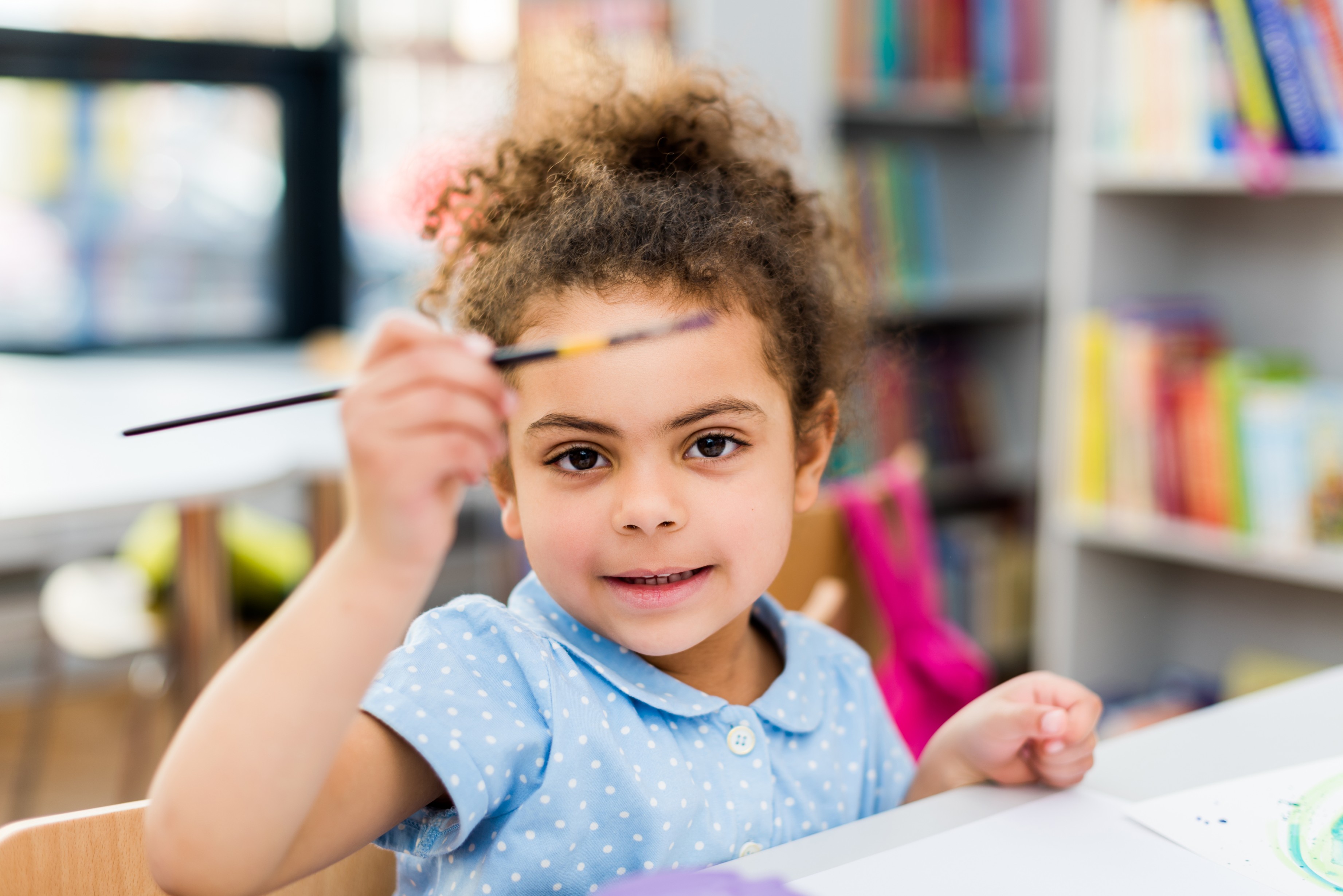 A young girl holding a pencil and looking at the camera while doing schoolwork.
