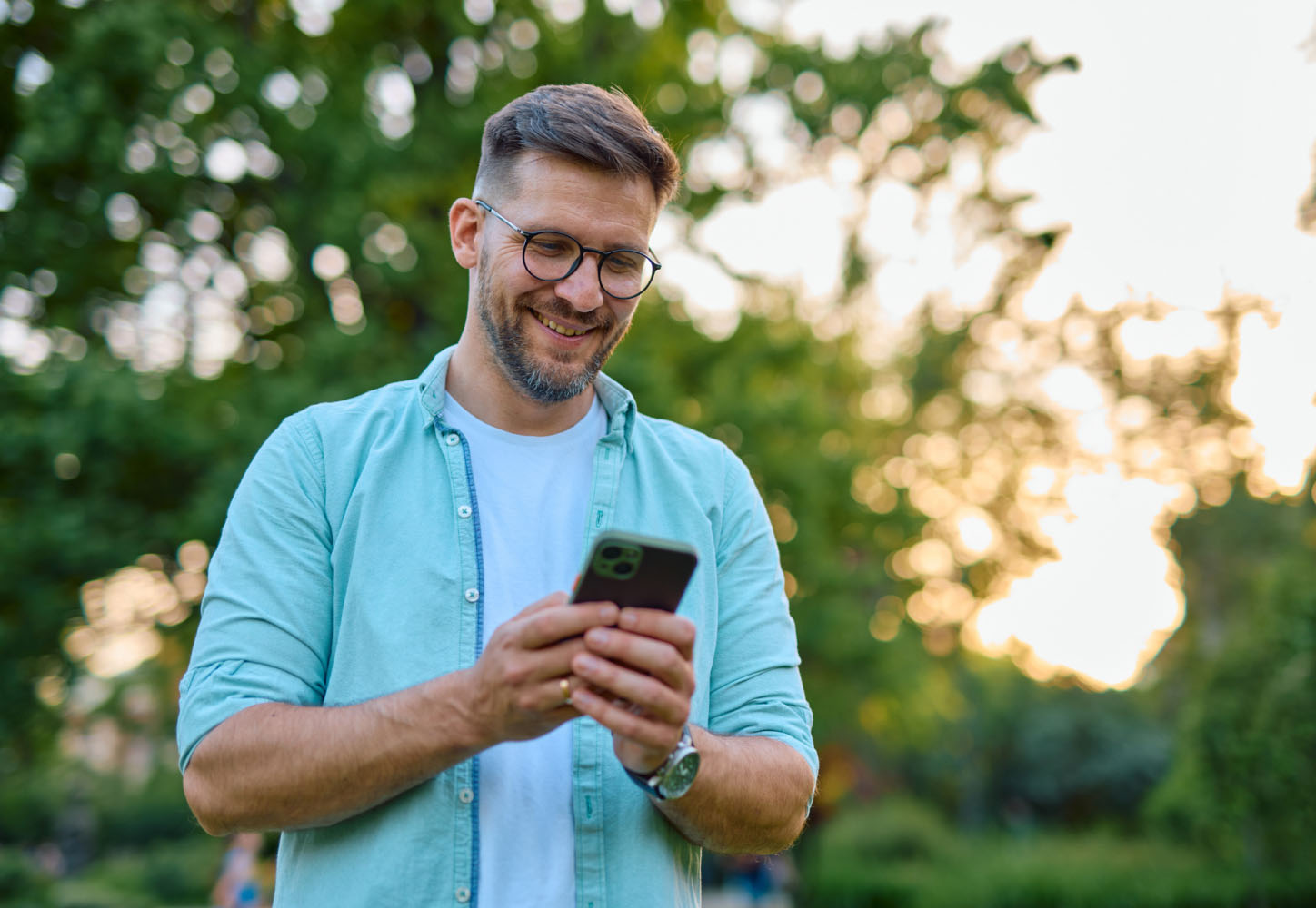A smiling man stands outside, looking at his phone.