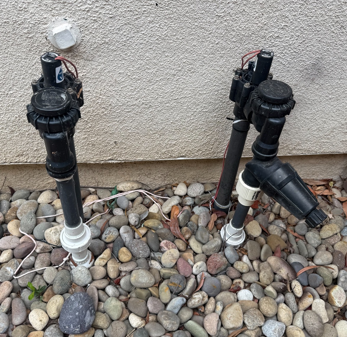 Sprinkler system valves and backflow risers against a stucco wall in a river-rock bed