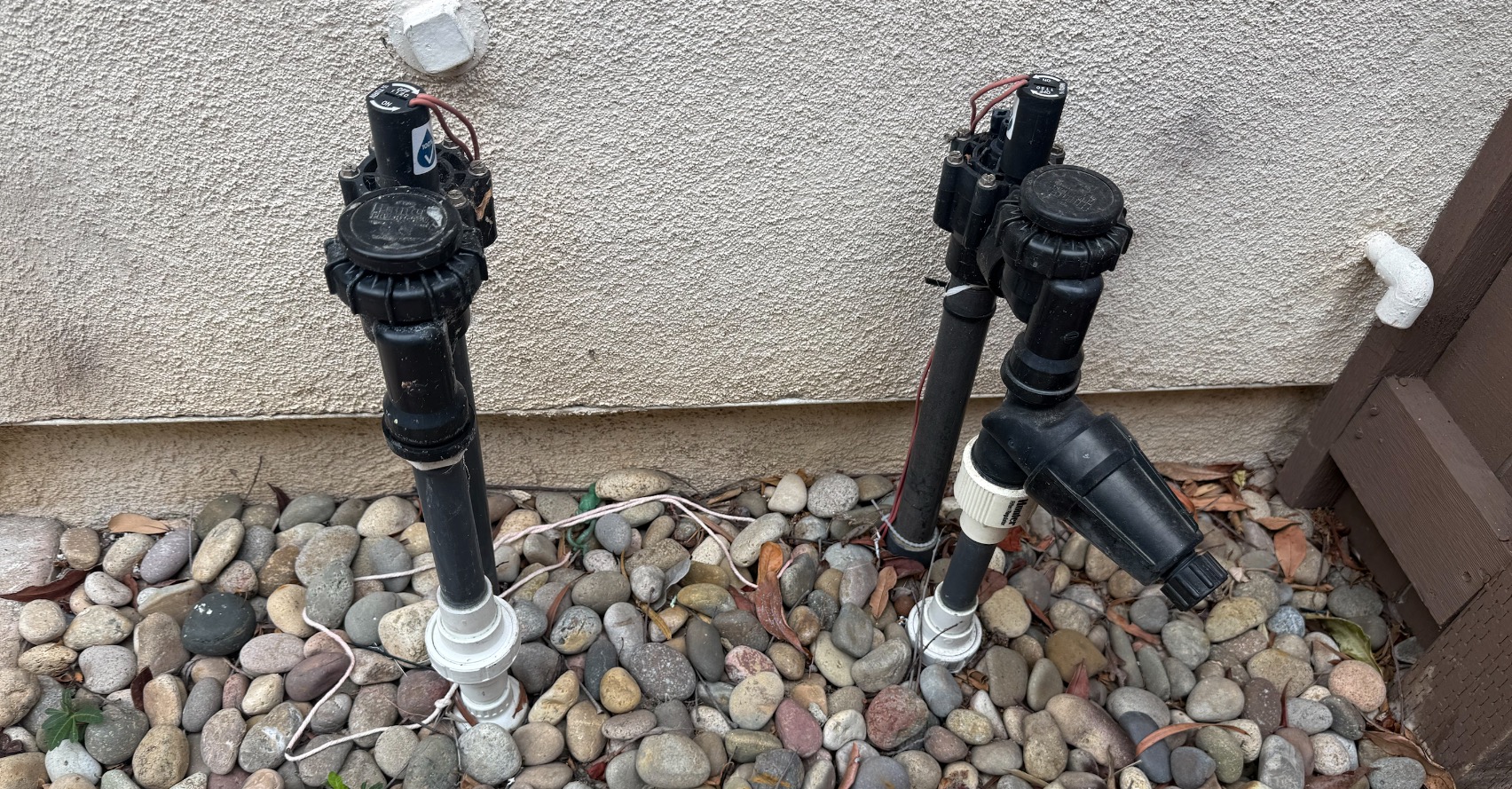 Sprinkler system valves and backflow risers against a stucco wall in a river-rock bed
