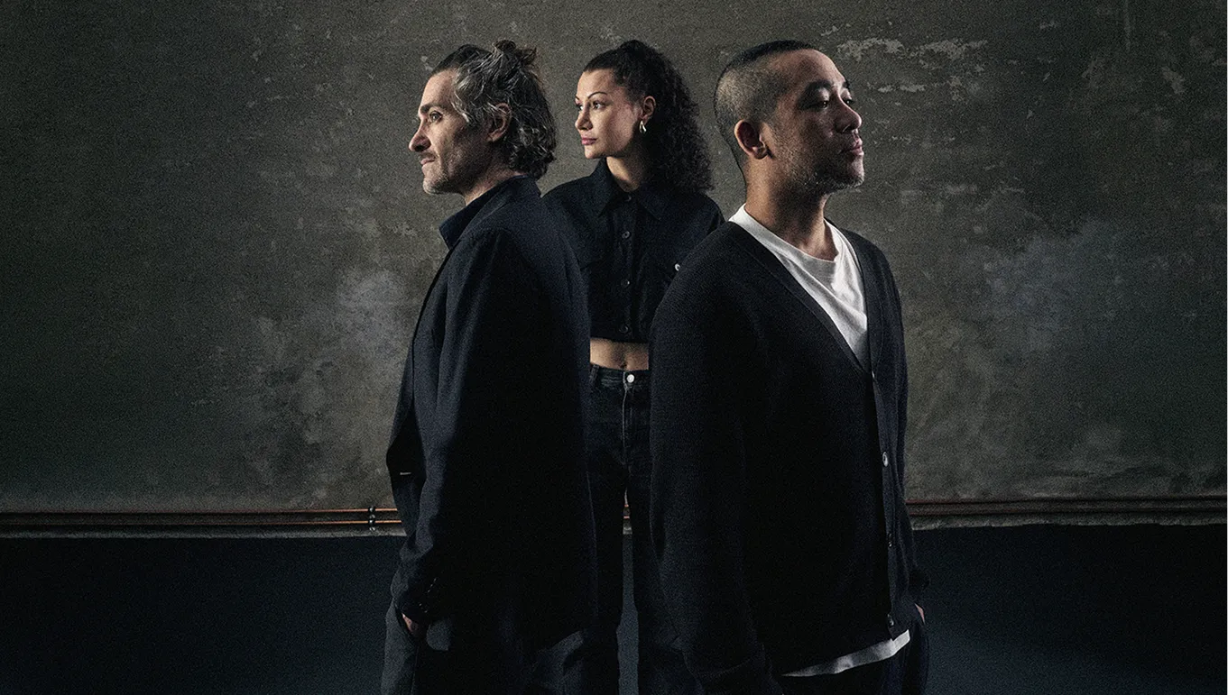 Die drei Gründer von Hot Studio – Sacha, Jasmin und Harold – stehen nebeneinander vor einem dunklen Studiohintergrund, alle in Schwarz gekleidet, mit Blick zur Seite.