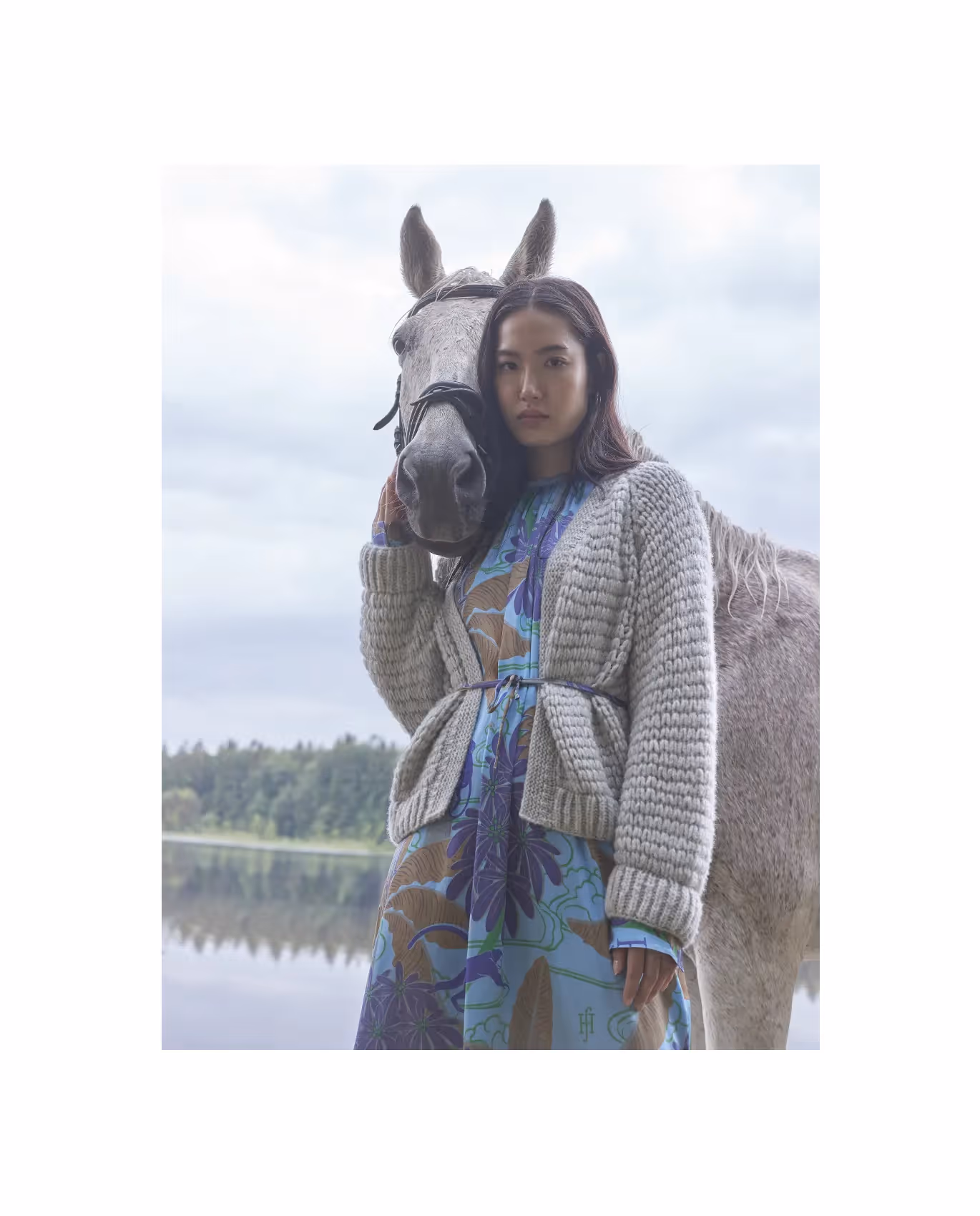 Frau in gemustertem Kleid und Strickjacke steht neben einem Pferd vor einem See