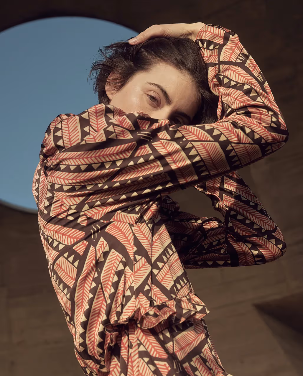 Frau trägt ein gemustertes Kleid in Rot- und Brauntönen und hält ihren Arm vor das Gesicht