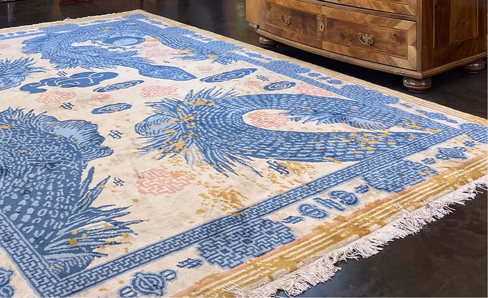 Teppich mit blauem Muster und Fransen liegt auf dunklem Boden neben einem Holzmöbel