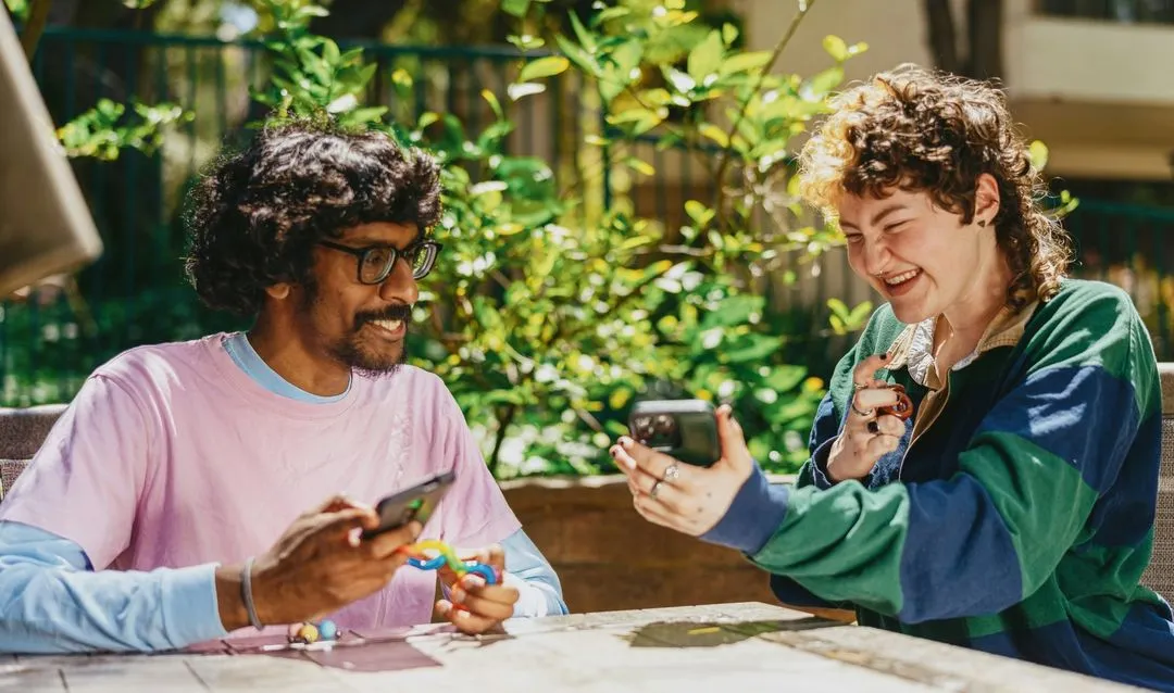 Two people sit outdoors at a table amidst lush greenery, smiling and looking at their phones. The scene conveys a joyful and relaxed atmosphere.
