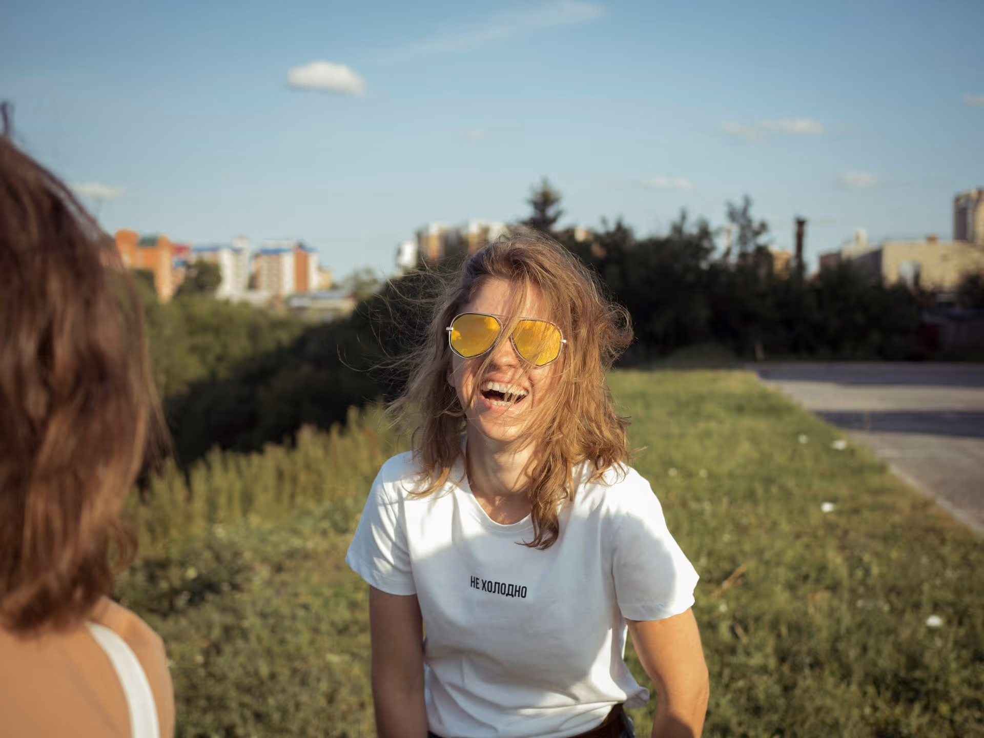 Young woman with wavy hair and yellow sunglasses, laughing joyfully outside. She's wearing a white T-shirt, green grass, and cityscape in the background.