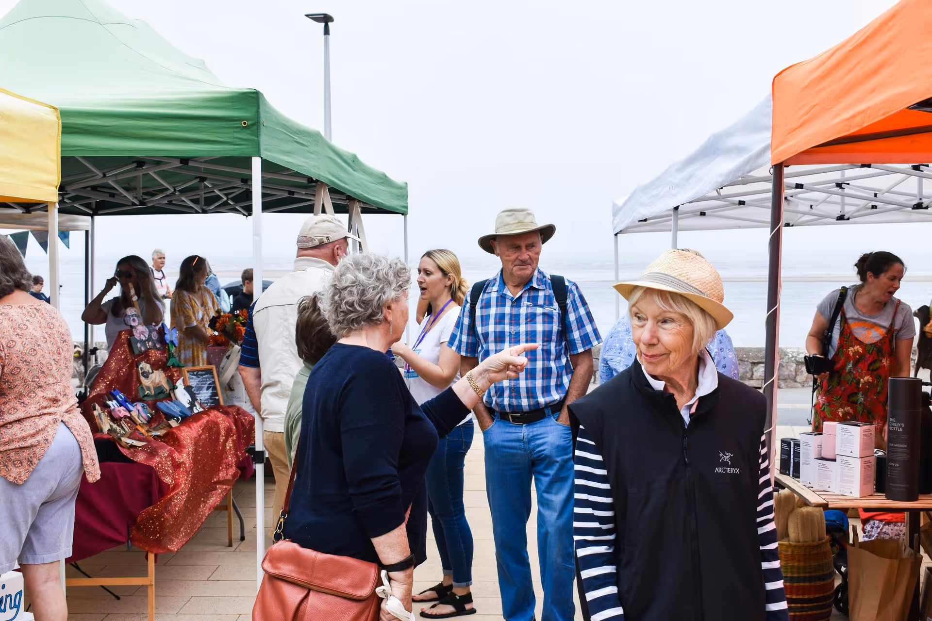 People enjoying a lively outdoor market by the sea, chatting and browsing stalls under colorful tents on a cloudy day, creating a vibrant atmosphere.