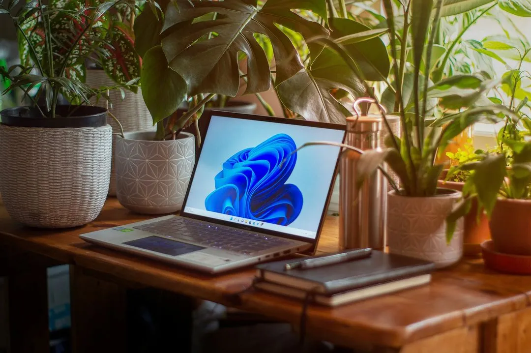 A cozy workspace with a laptop displaying a blue abstract screen, surrounded by lush green plants. Notebooks and a pen add a productive vibe.