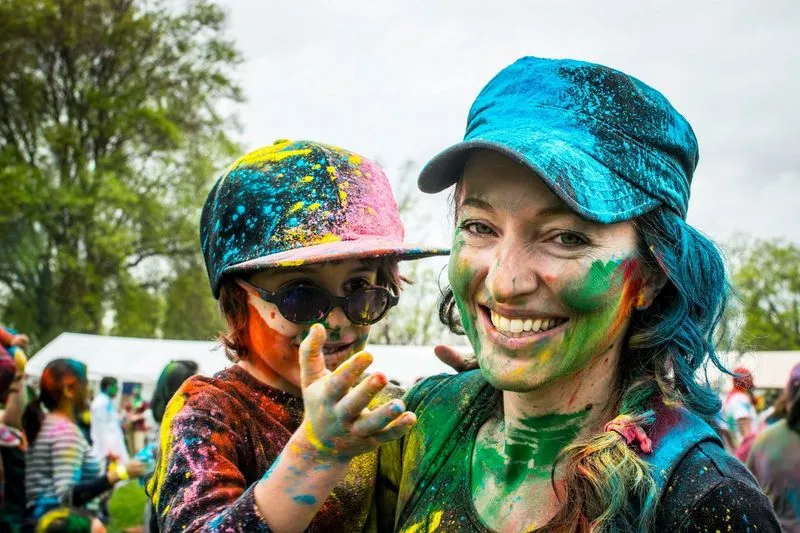 A woman and child, covered in vibrant colored powders, smile joyfully at a festival. They wear colorful caps and the background shows people and trees.