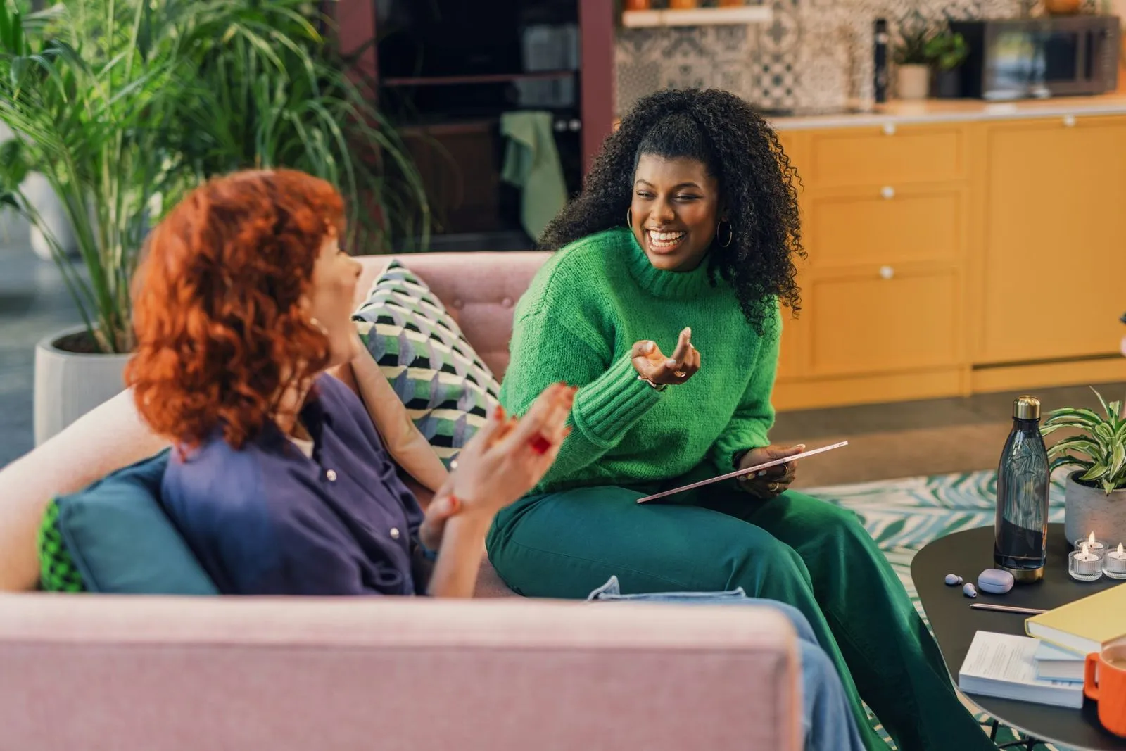 Two women sit on a sofa in a cozy room, engaged in a lively conversation. One wears a green sweater, the other a blue shirt. Both are smiling warmly.