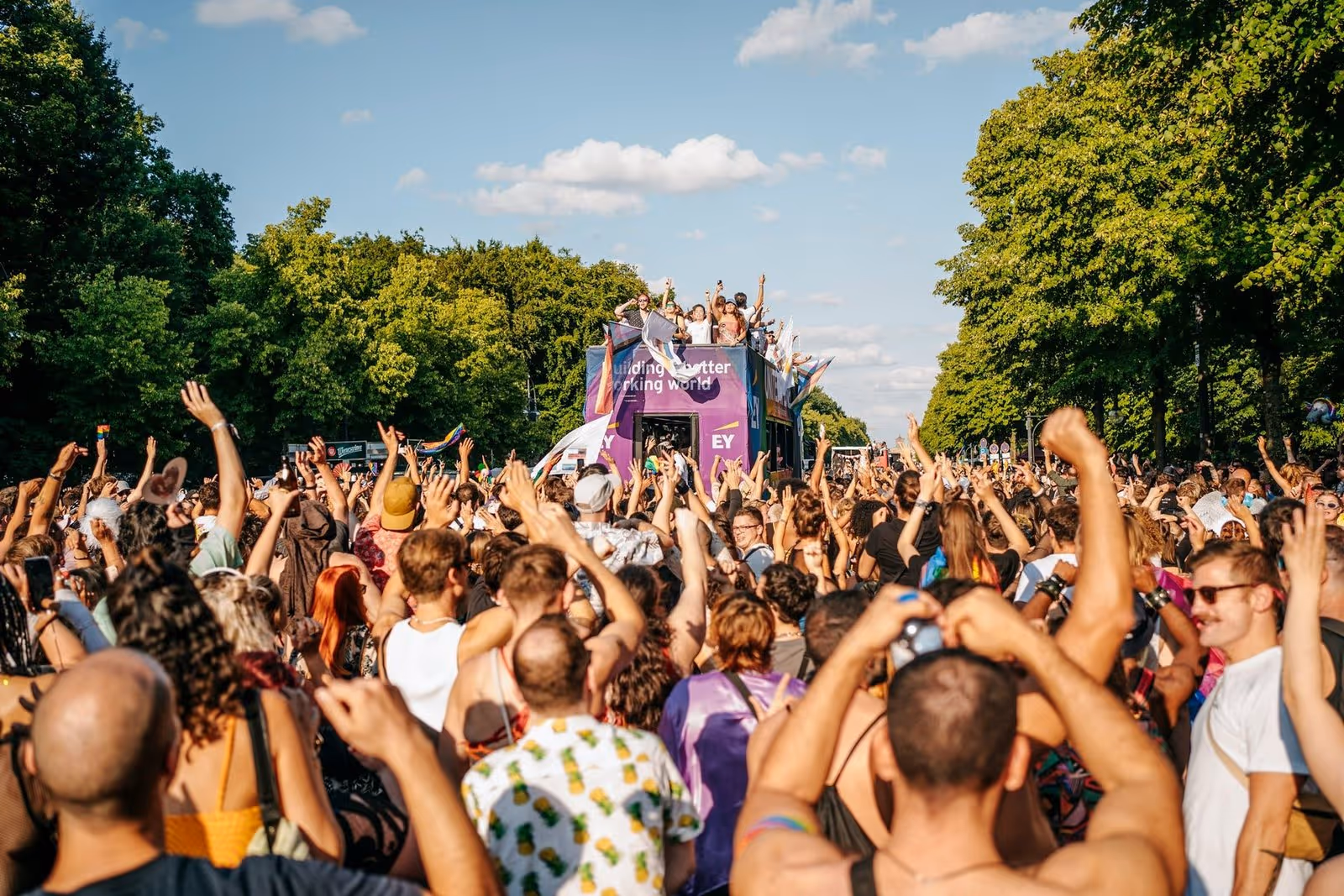 A lively crowd celebrates at a Pride parade under a sunny sky. People cheer and wave flags near a colorful float surrounded by lush green trees.