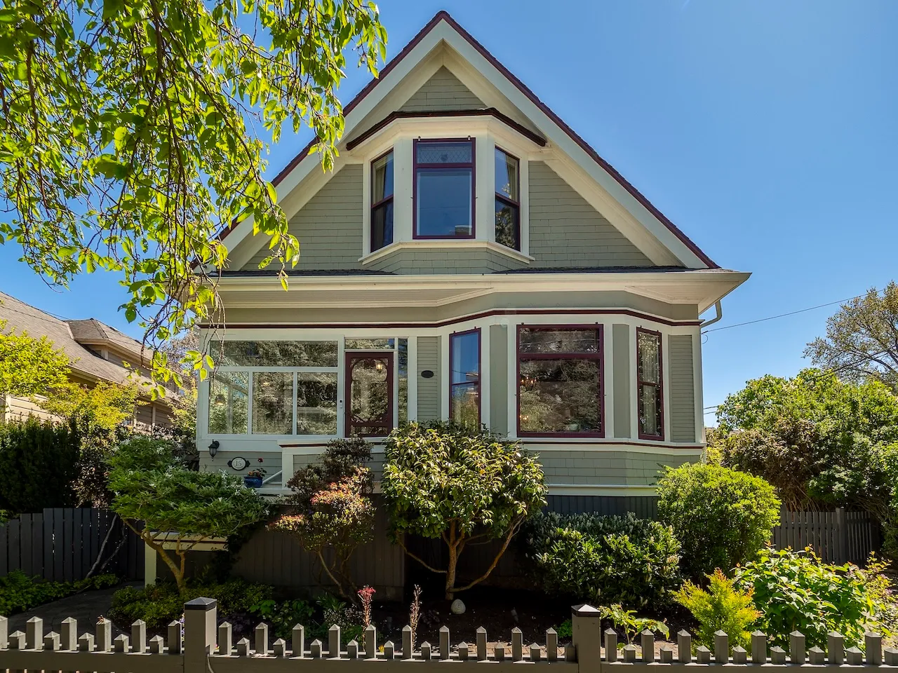 Two-story house with light green siding, white trim, burgundy window frames, front porch, and lush greenery under a clear blue sky. Berk Christensen Real Estate Victoria BC.