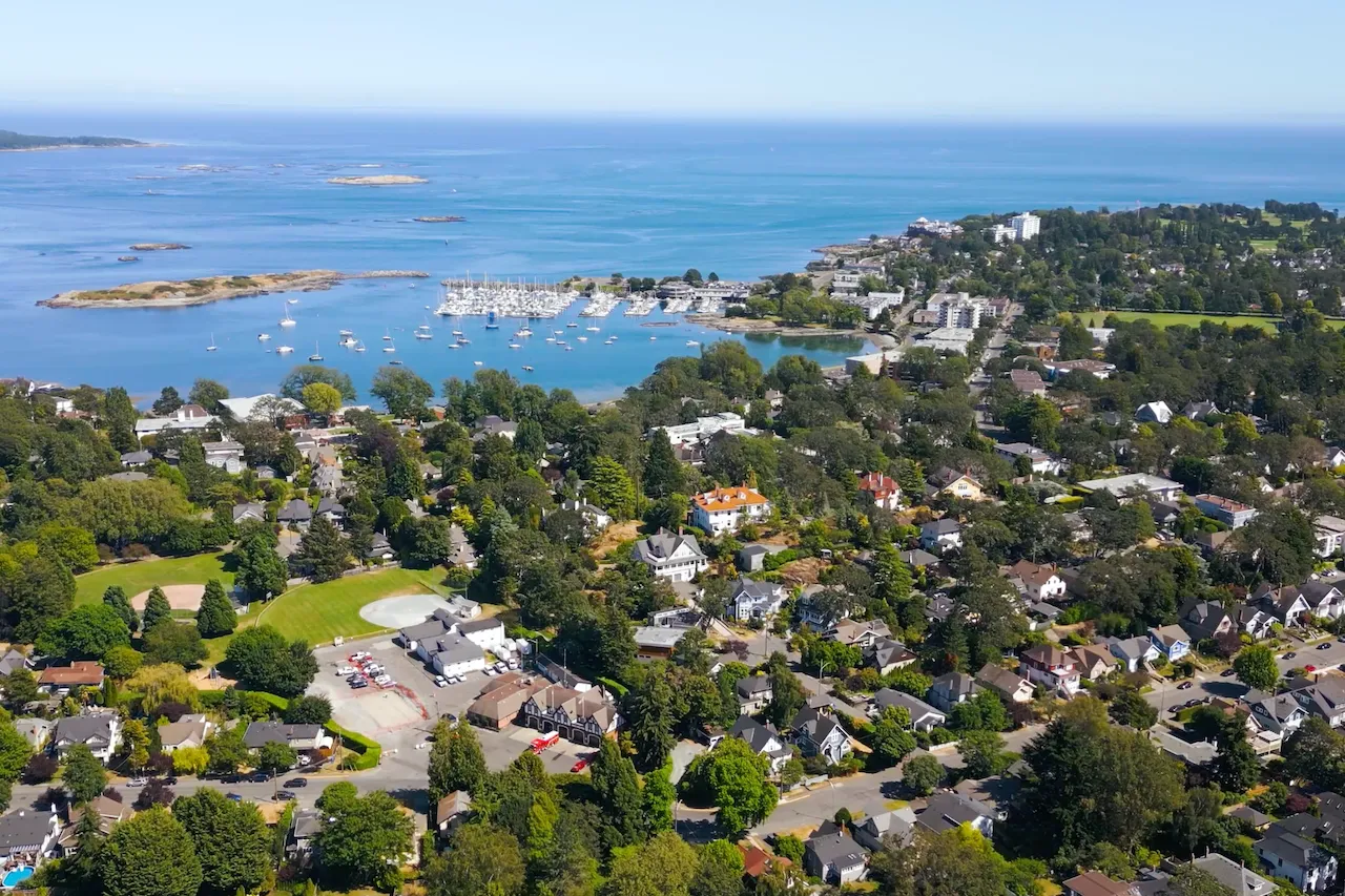 Aerial view of a coastal town with green trees, residential houses, a marina with boats, and calm blue sea under clear sky. Berk Christensen Real Estate Victoria BC.