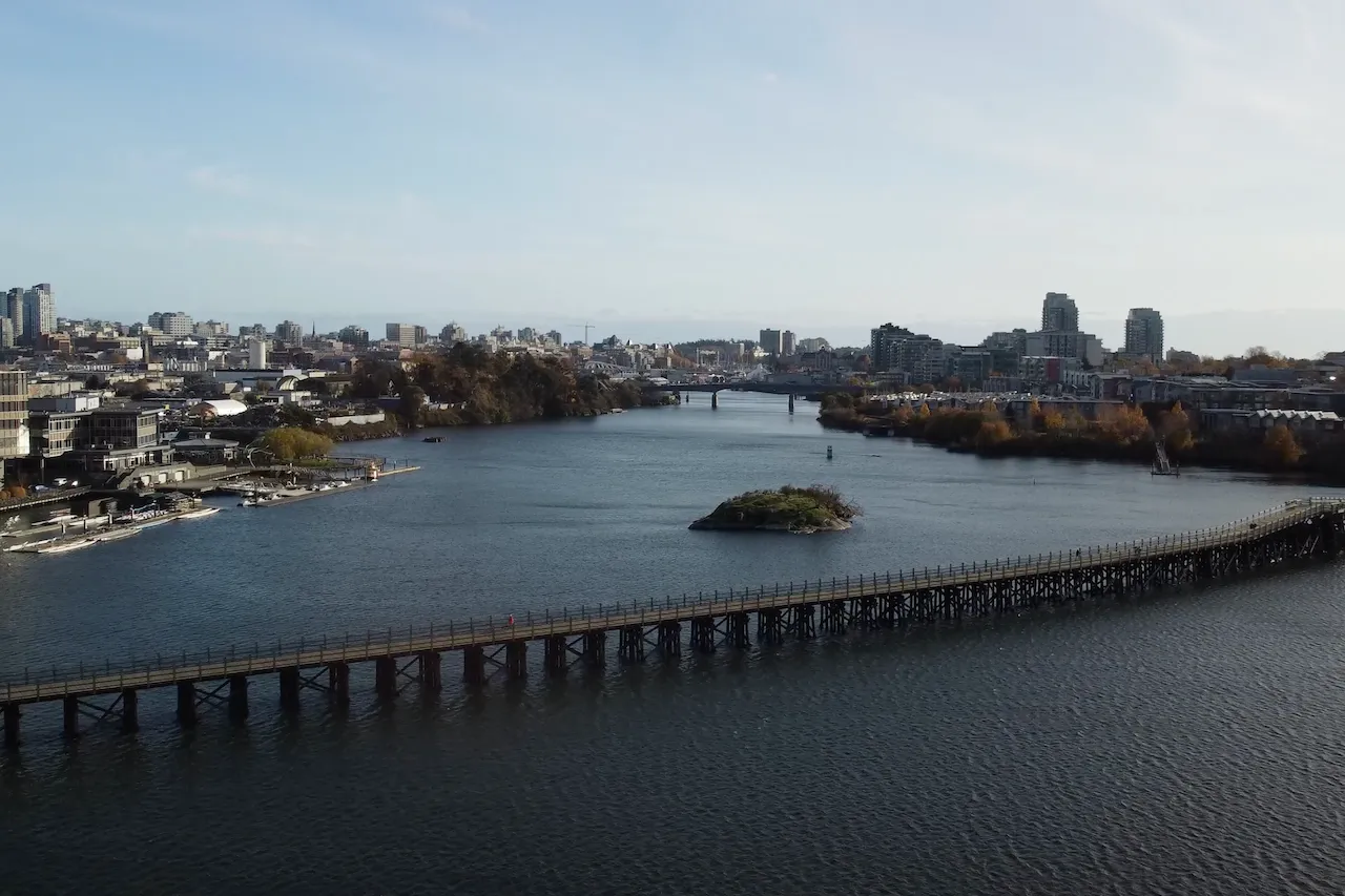 Wooden bridge crossing a wide river with a small island in the middle and a city skyline in the background under a clear sky. Berk Christensen Real Estate Victoria BC.