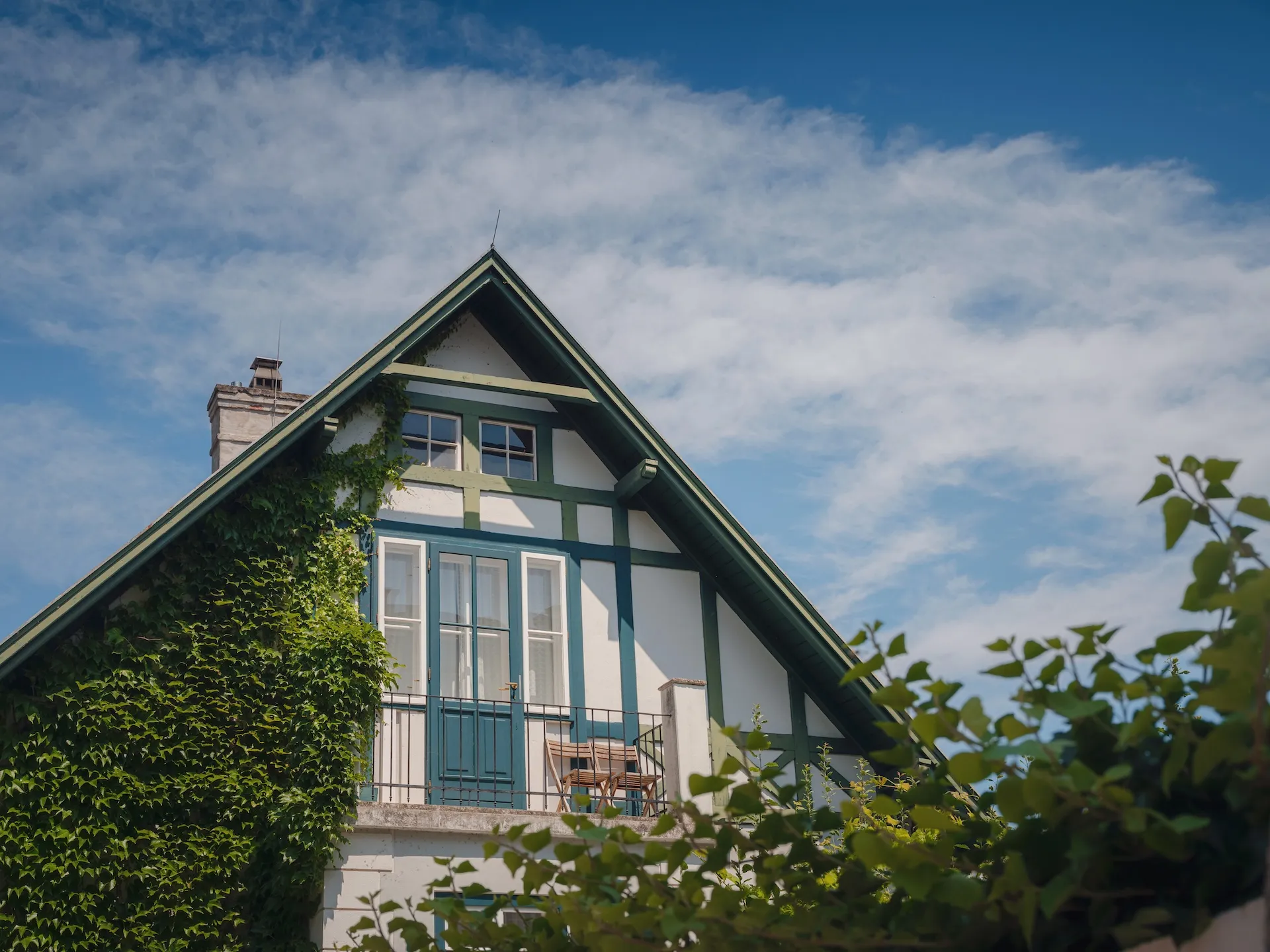 Upper section of a white house with green trim, a balcony with wooden chairs, and ivy climbing the wall under a partly cloudy blue sky. Berk Christensen Real Estate Victoria BC.