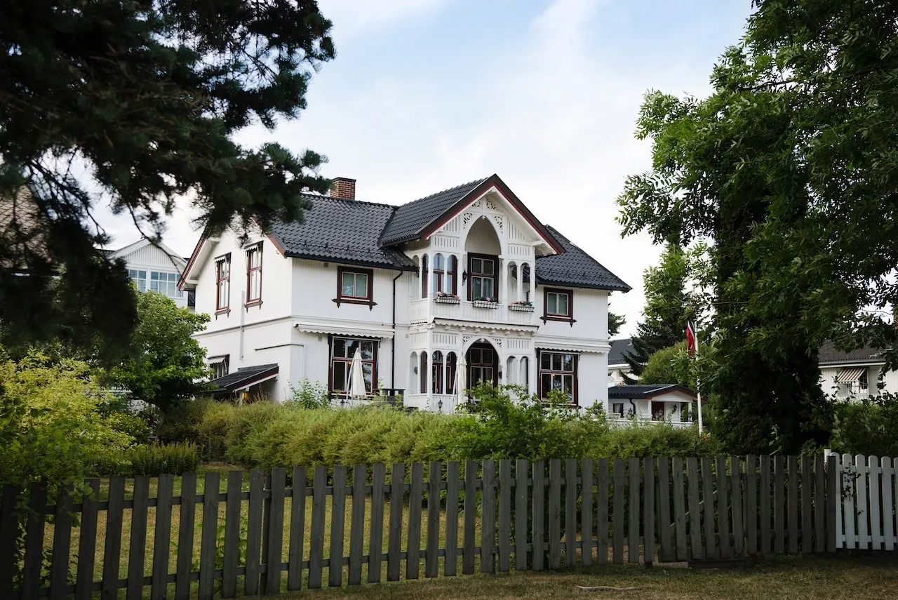 White two-story house with ornate wooden balcony, surrounded by green bushes, large trees, and a wooden fence. Berk Christensen Real Estate Victoria BC.