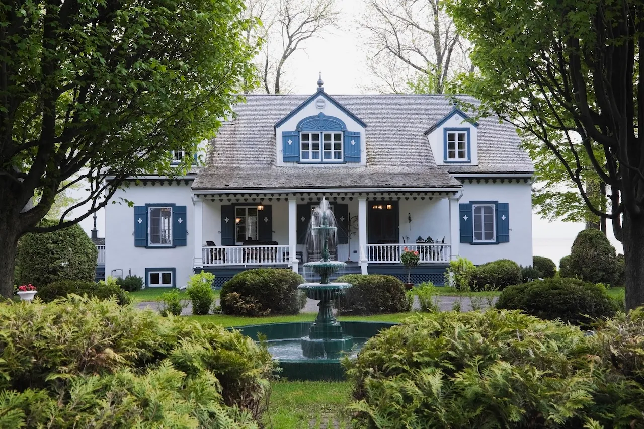 White cottage-style house with blue trim, a front porch, and a central green water fountain surrounded by green bushes and trees. Berk Christensen Real Estate Victoria BC.