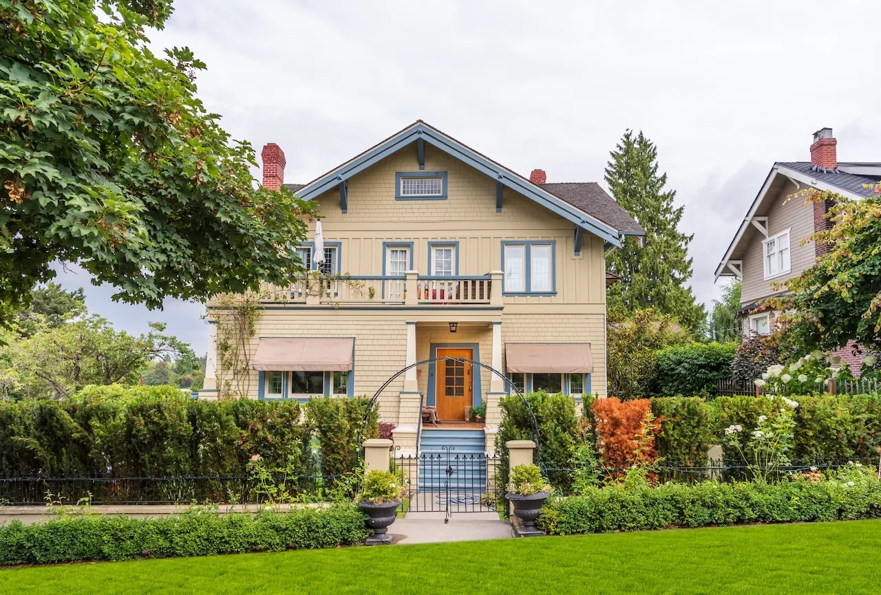 Two-story beige house with blue trim, front garden with hedge, green lawn, and gated entrance. Berk Christensen Real Estate Victoria BC.