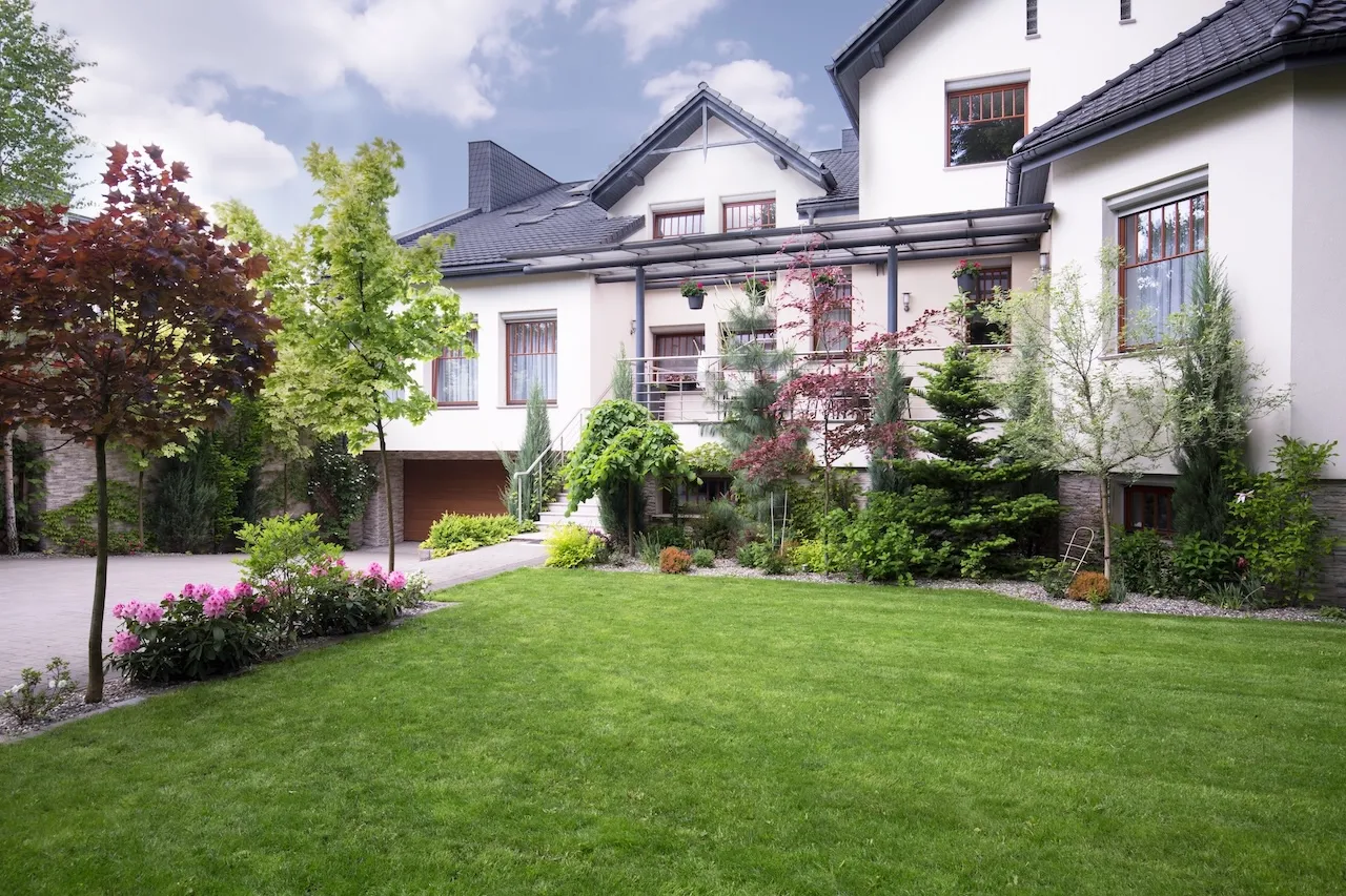 Modern two-story white house with dark roof, wooden windows, and a well-maintained garden with various trees and green lawn. Berk Christensen Real Estate Victoria BC.