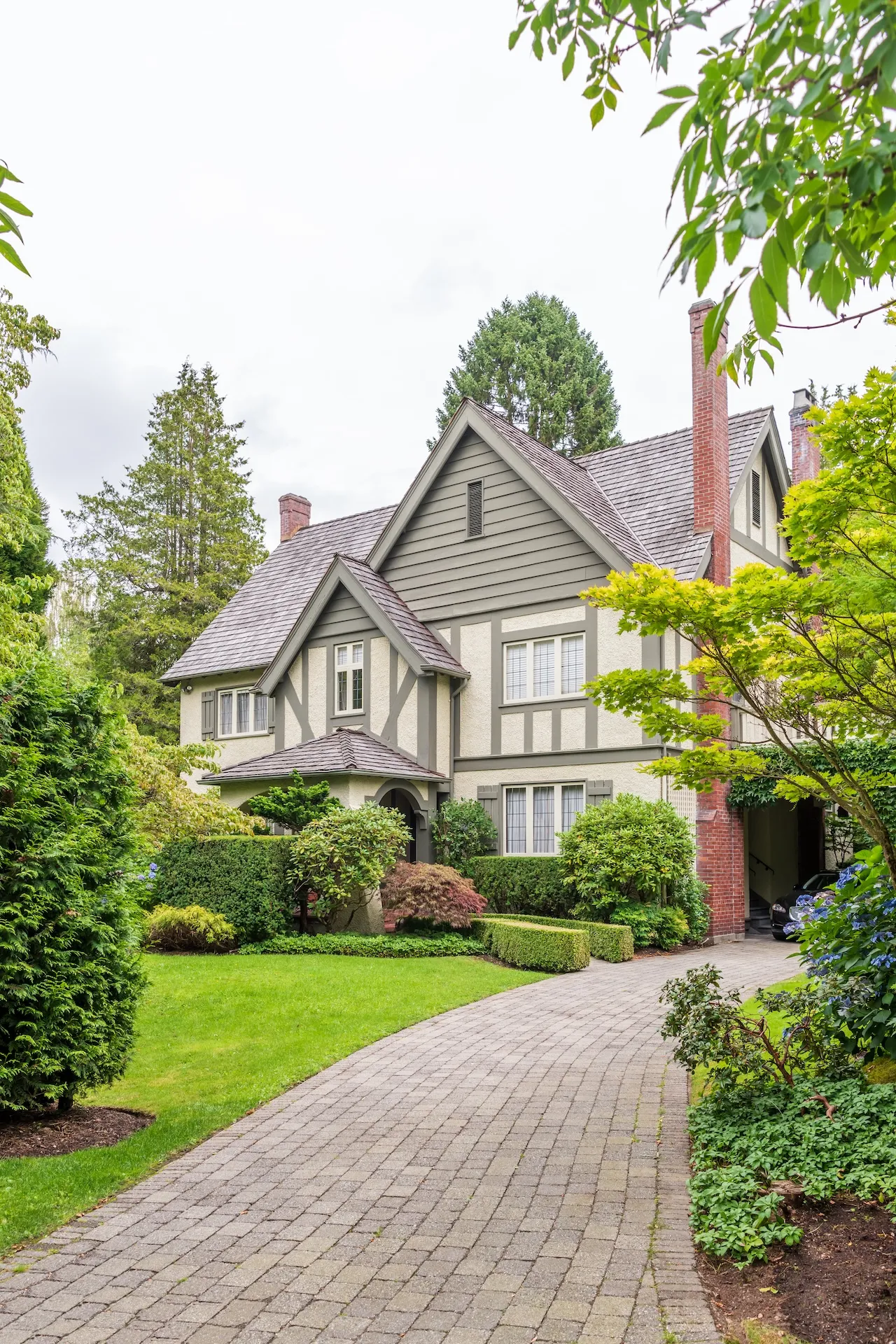 Tudor-style house with gray siding, brick chimneys, and a curved paved driveway surrounded by green trees and bushes. Berk Christensen Real Estate Victoria BC.