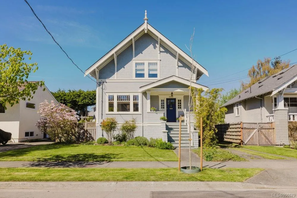 Two-story gray house with white trim, a blue front door, and a well-maintained lawn with small trees and shrubs under a clear blue sky. Berk Christensen Real Estate Victoria BC.
