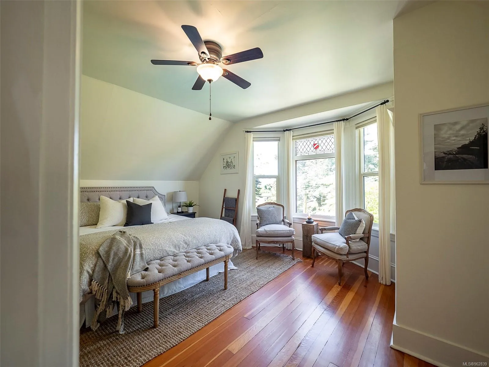 Bright bedroom with a large bed, cushioned bench, two armchairs near a bay window, and a ceiling fan overhead. Berk Christensen Real Estate Victoria BC.