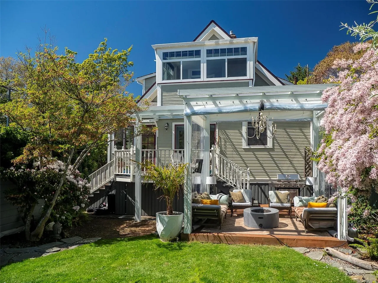 Backyard patio with cushioned seating, a fire pit, and a transparent pergola attached to a two-story house with greenery and flowering trees. Berk Christensen Real Estate Victoria BC.