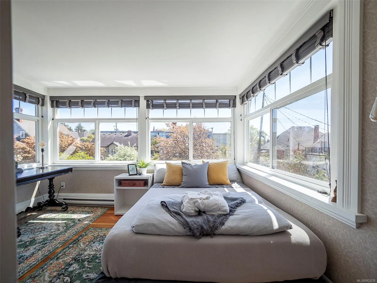 Bright bedroom with large windows, a bed with three pillows, a small white bedside table, and a patterned rug on wooden floor. Berk Christensen Real Estate Victoria BC.