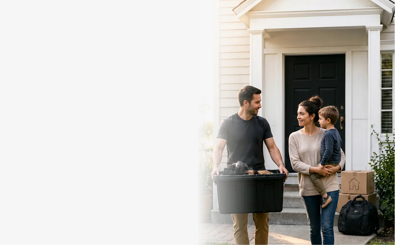 Couple with a child moving into a house, carrying a storage bin and standing near boxes by the door.