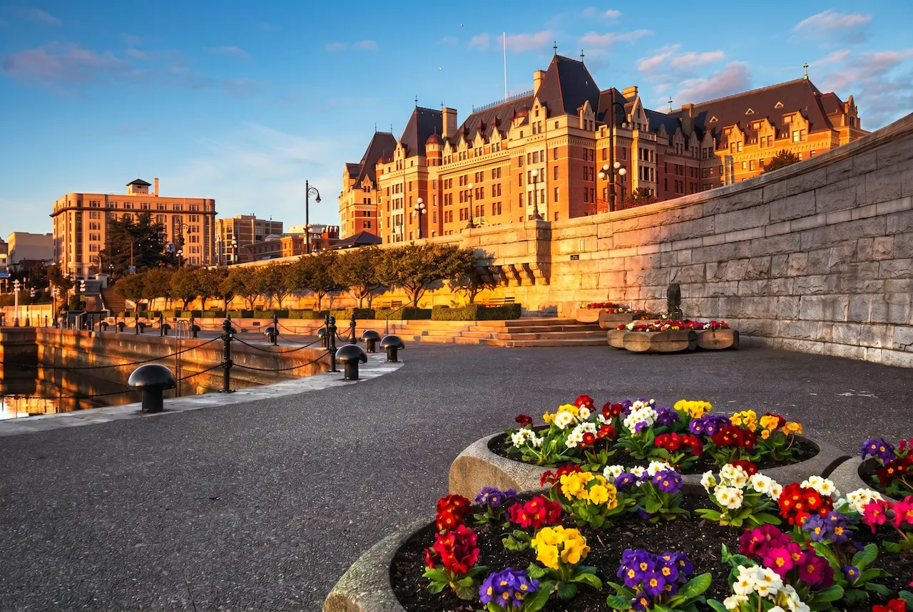 Sunlit historic building with dark roofs near a waterfront path and colorful flower beds in the foreground.