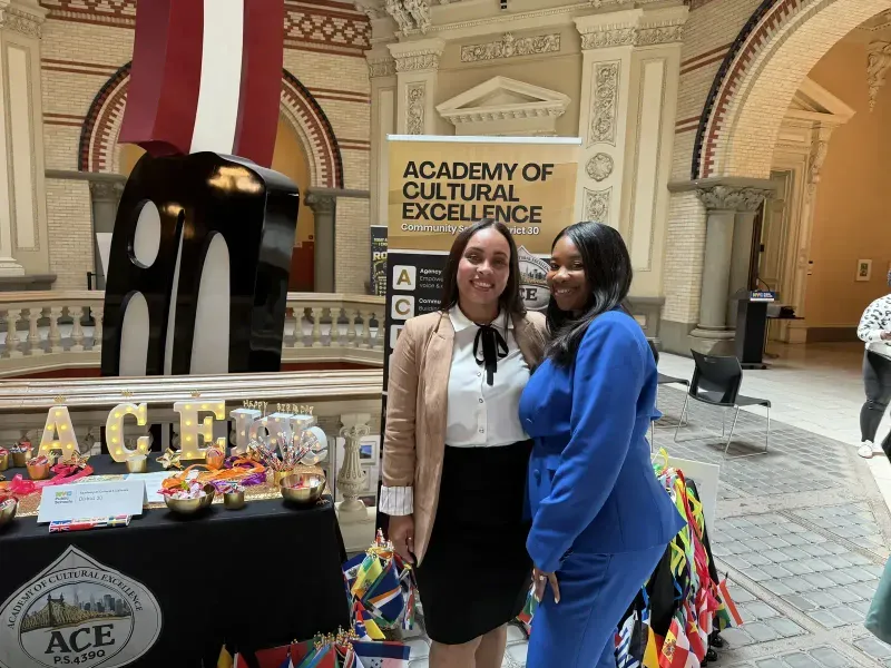 Two ACE educators pose together next to the Academy of Cultural Excellence banner