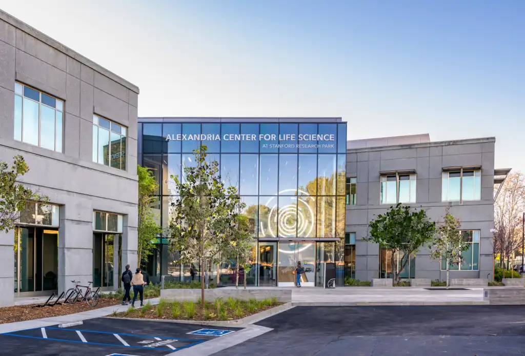 Modern campus building with glass facade labeled Alexandria Center for Life Science at Stanford Research Park, with trees and people outside.