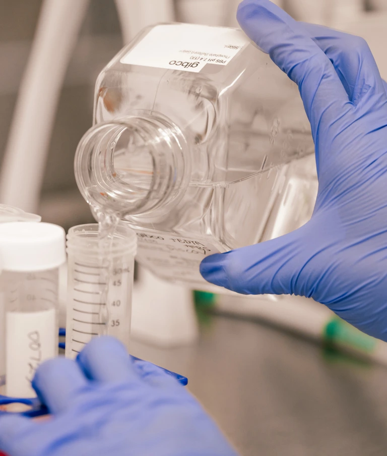 Gloved hands pouring liquid from a clear square bottle into a graduated plastic test tube in a laboratory setting.