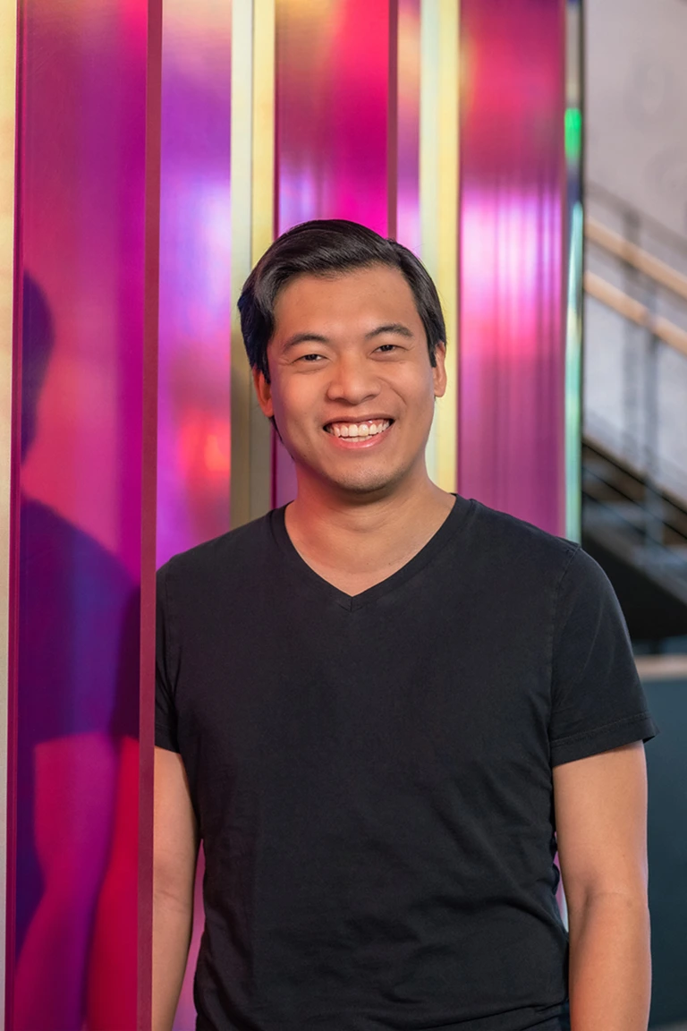 Smiling man wearing a black V-neck T-shirt standing in front of a colorful pink and purple wall with gold vertical lines.