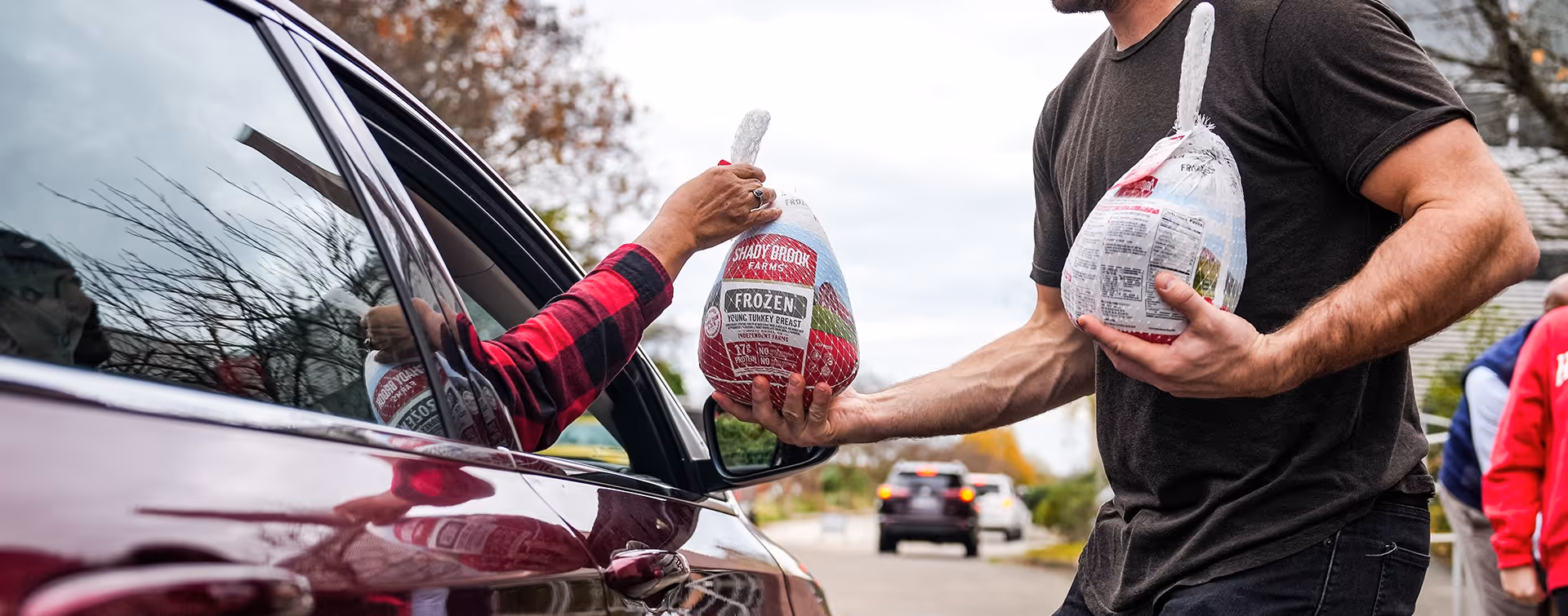 Person handing a packaged frozen turkey to a women in a car at a drive through food pantry
