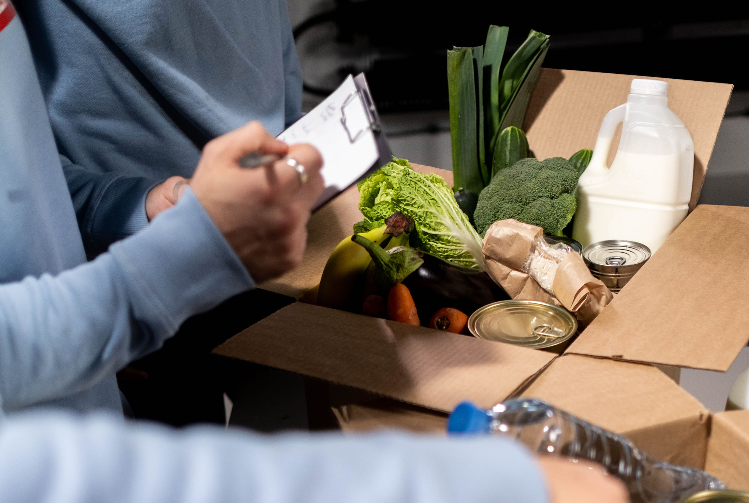 Person in blue sweater holding a clipboard checking a cardboard box filled with fresh vegetables, canned food, milk, and grocer items.