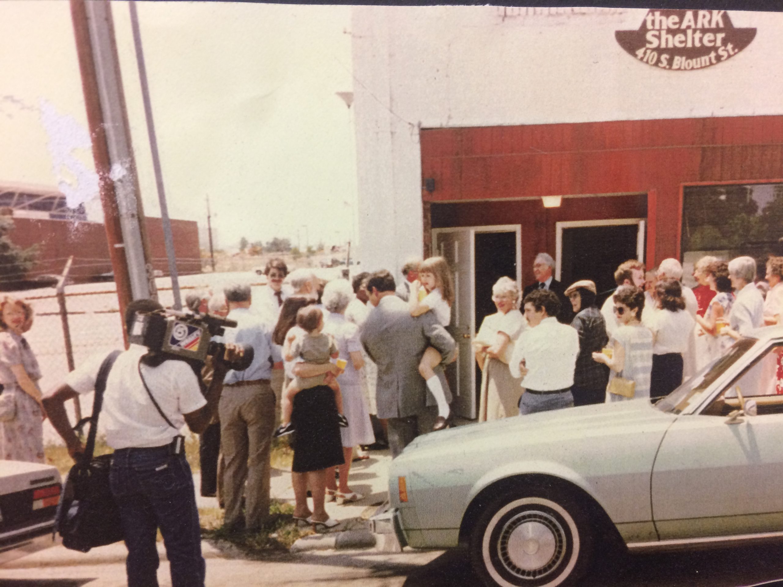 Crowd gathered in 1981 at the entrance of the old ARK Shelter, with a cameraman filming the event and a man holding a child entering the building.
